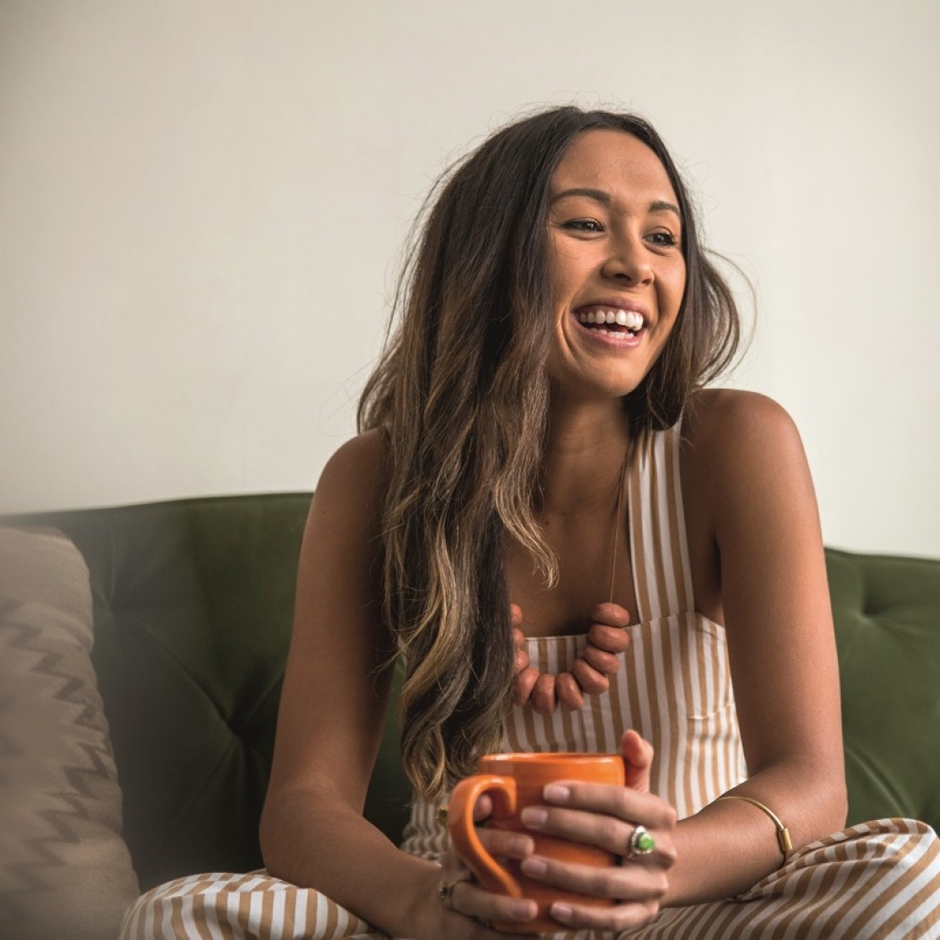 A young woman smiling whilst sat on a sofa and enjoying a cup of coffee