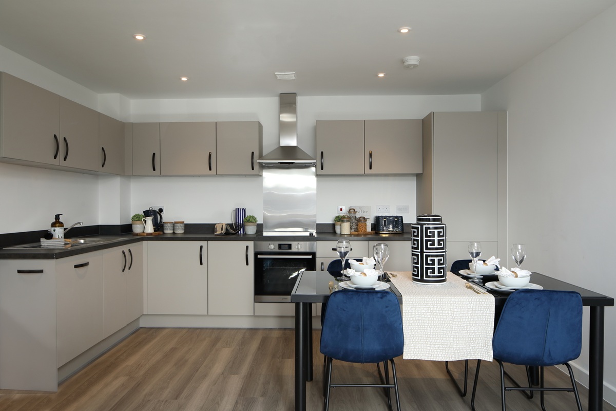 Modern open plan kitchen and dining area with grey cabinets and wooden floor, and a set dining table with blue chairs in a two bedroom apartment in Chapel House, Potter's Grange.