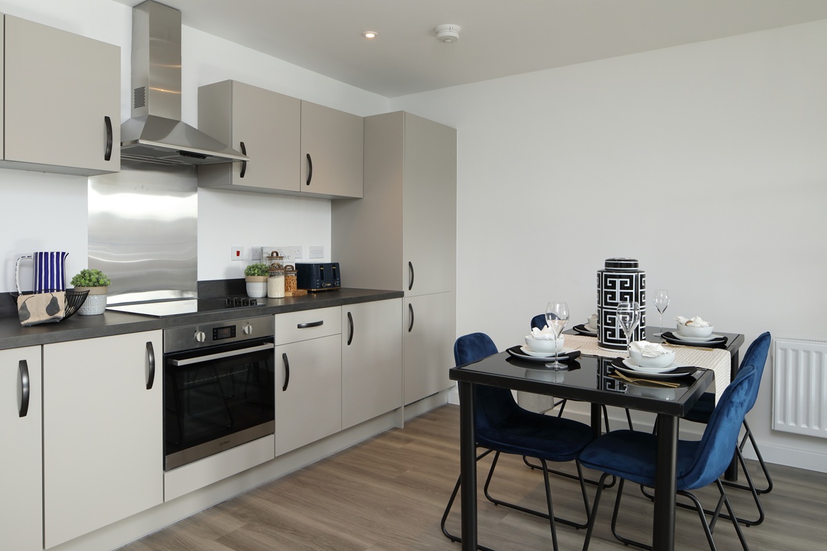Modern open plan kitchen and dining area with grey cabinets and wooden floor, and a set dining table with blue chairs in a two bedroom apartment in Chapel House, Potter's Grange.