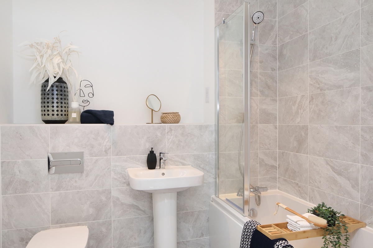A bathroom with a shower over bath, sink, and toilet with a chrome flush plate in a Chapel House apartment at Potter’s Grange in Ashby.