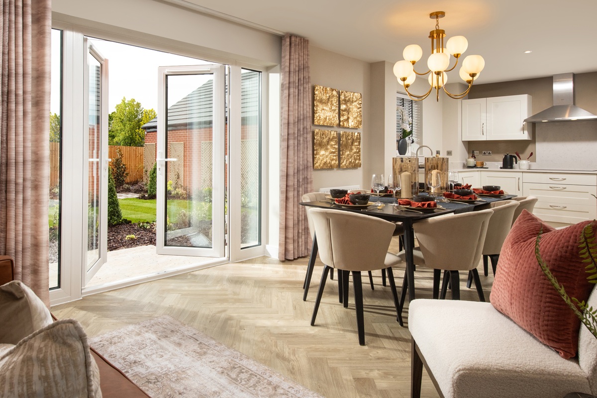 Open-plan kitchen and dining area in a Crest Nicholson home, featuring French doors to the garden, a black dining table with beige chairs, white cabinetry, and herringbone floor.