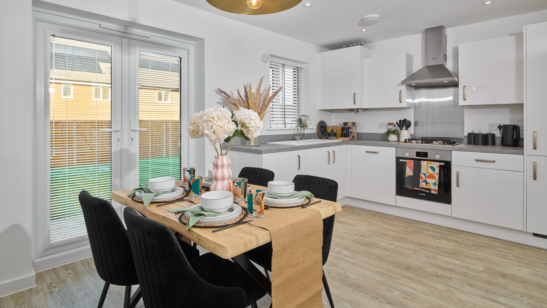 Open-plan kitchen and dining area in a Crest Nicholson home, featuring French doors to the garden, a wooden dining table with black chairs, white cabinetry, and a grey worktop.
