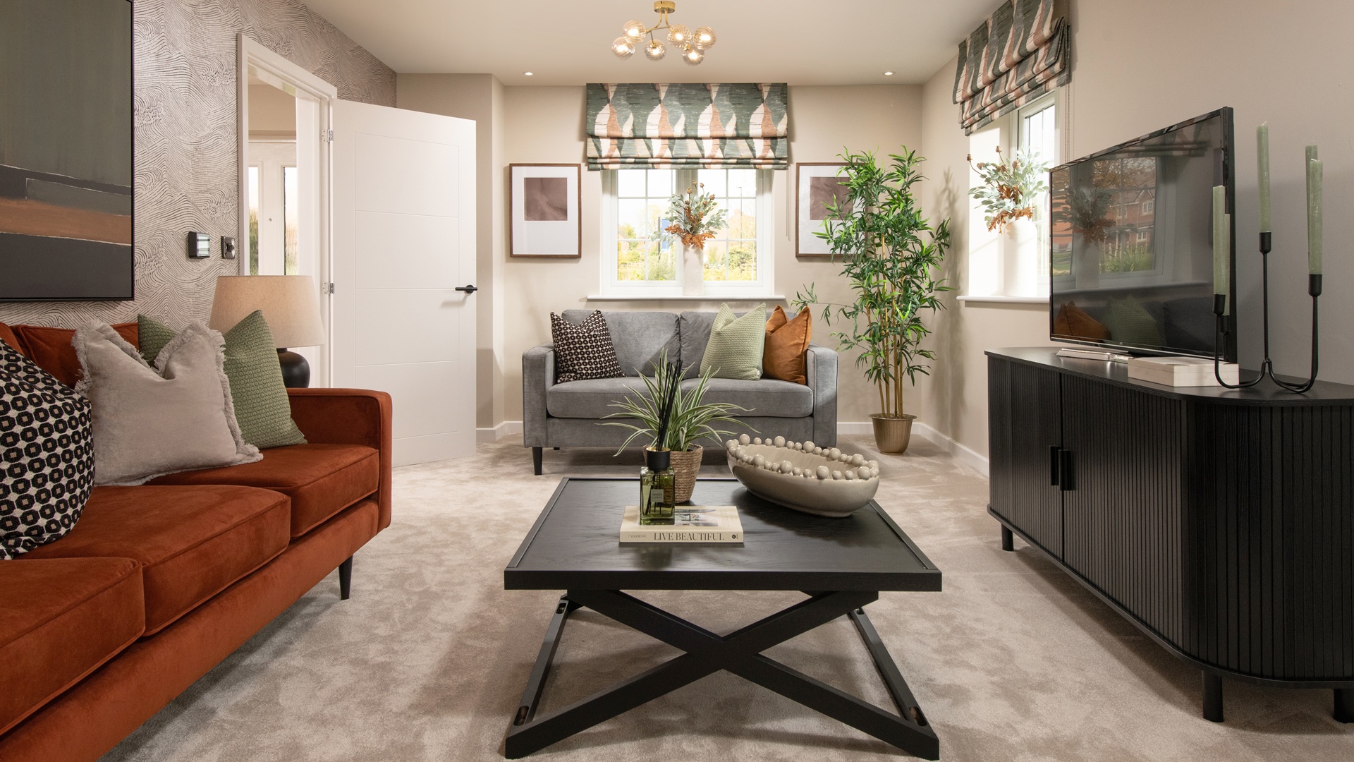 A cosy living room featuring a rust-coloured sofa on the left, a grey sofa in the background, a dark wood coffee table with an X-base, a dark television console, and a large potted plant near a window in a Marlborough Crest Nicholson home.